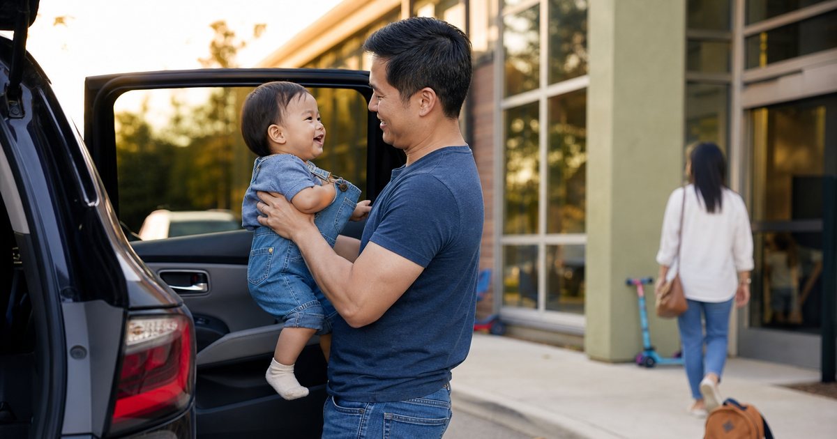 Father lifting a toddler into a car seat outside a daycare building at pickup, a small backpack on the sidewalk, another parent walking toward the entrance in the background, late afternoon