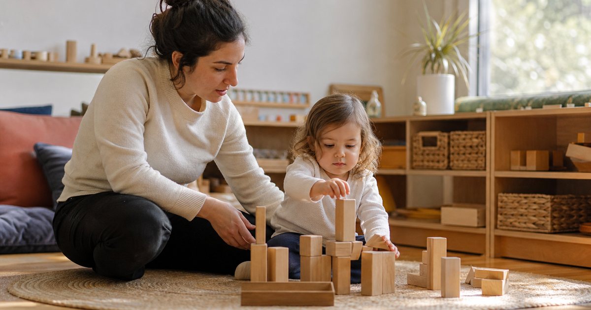A preschool teacher kneeling on a low rug beside a toddler stacking wooden blocks in a sunlit classroom, soft cushions and a low shelf of woven baskets behind them, warm afternoon light through a tall window