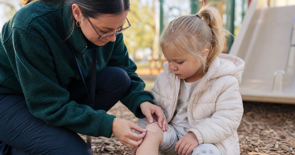 Daycare teacher kneeling beside a toddler on a playground, gently applying a bandage to a scraped knee, wood chips and a low slide in the background, afternoon sun