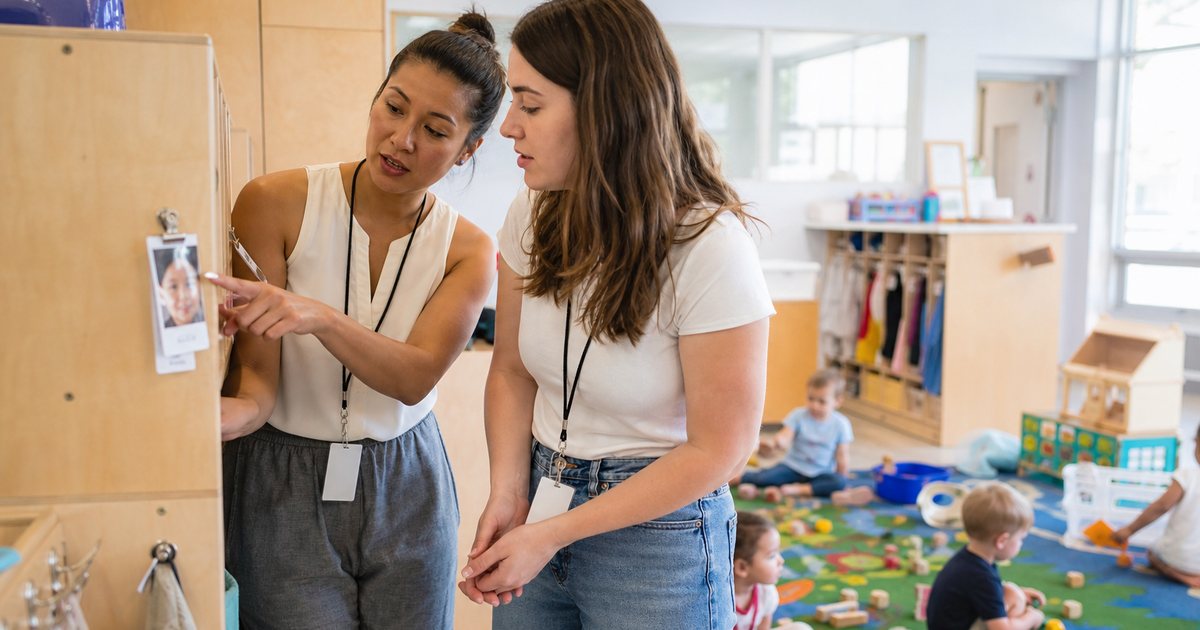 A new teacher in a daycare classroom reading a laminated card clipped to a cubby while toddlers play with blocks on a rug nearby, morning light, a lead teacher pointing at the card